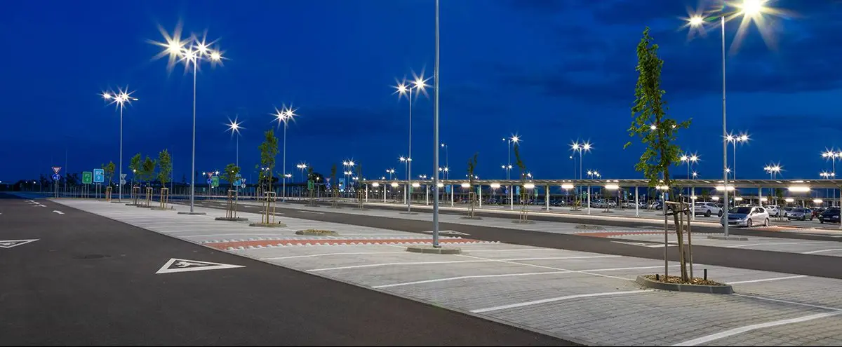 Well-lit parking lot at dusk with bright LED street lights, trees, and parked cars