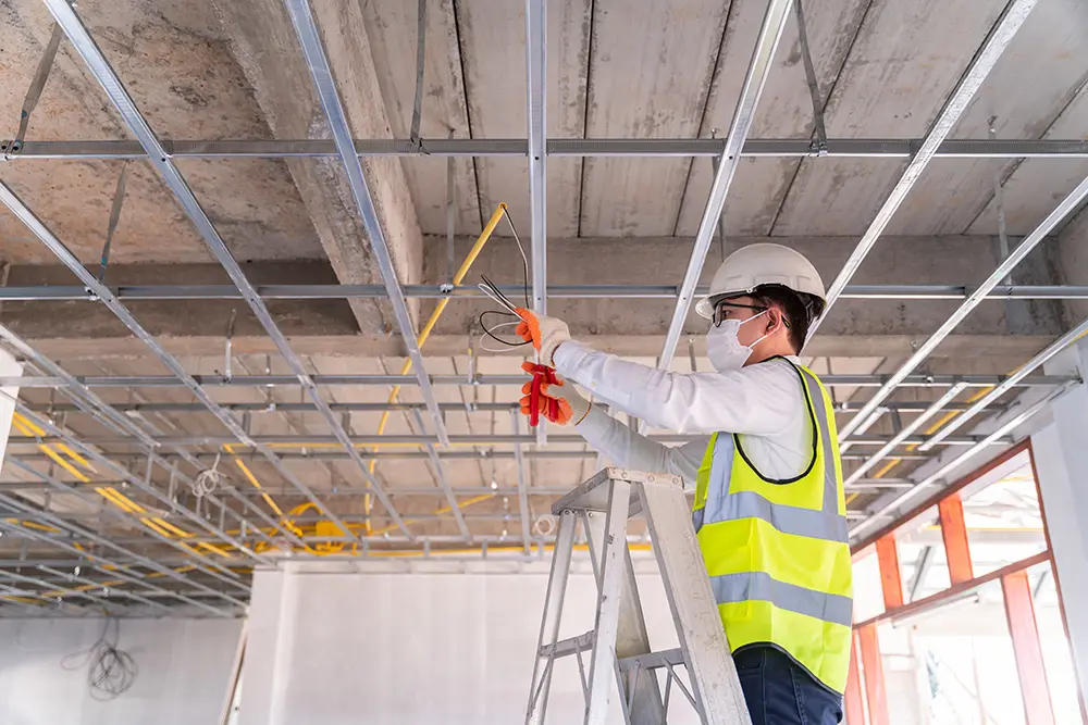 Electrician in white shirt and hard hat installing wiring on ceiling framework